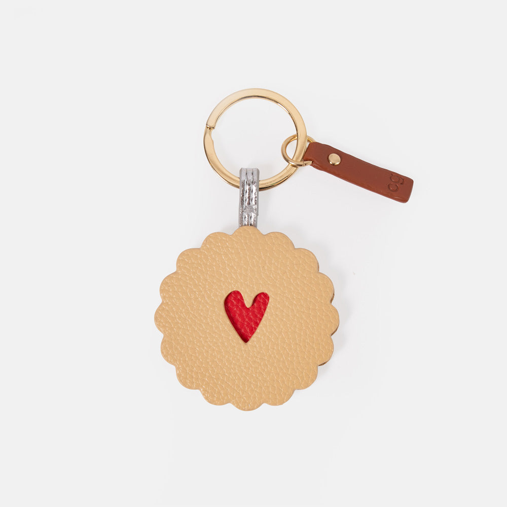 A round, scalloped-edge leather keyring charm designed like a jammy dodger biscuit, featuring a textured tan body with a small red heart in the center. It is attached to a gold-tone split ring and a brown leather tag with a gold rivet, set against a plain light gray background.