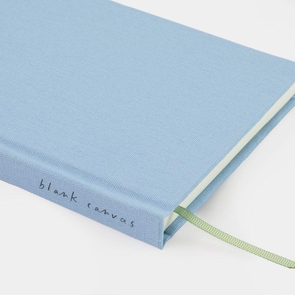 A light blue canvas-bound notebook with a green ribbon bookmark and the words 'blank canvas' printed on the spine, shown on a clean white background.
