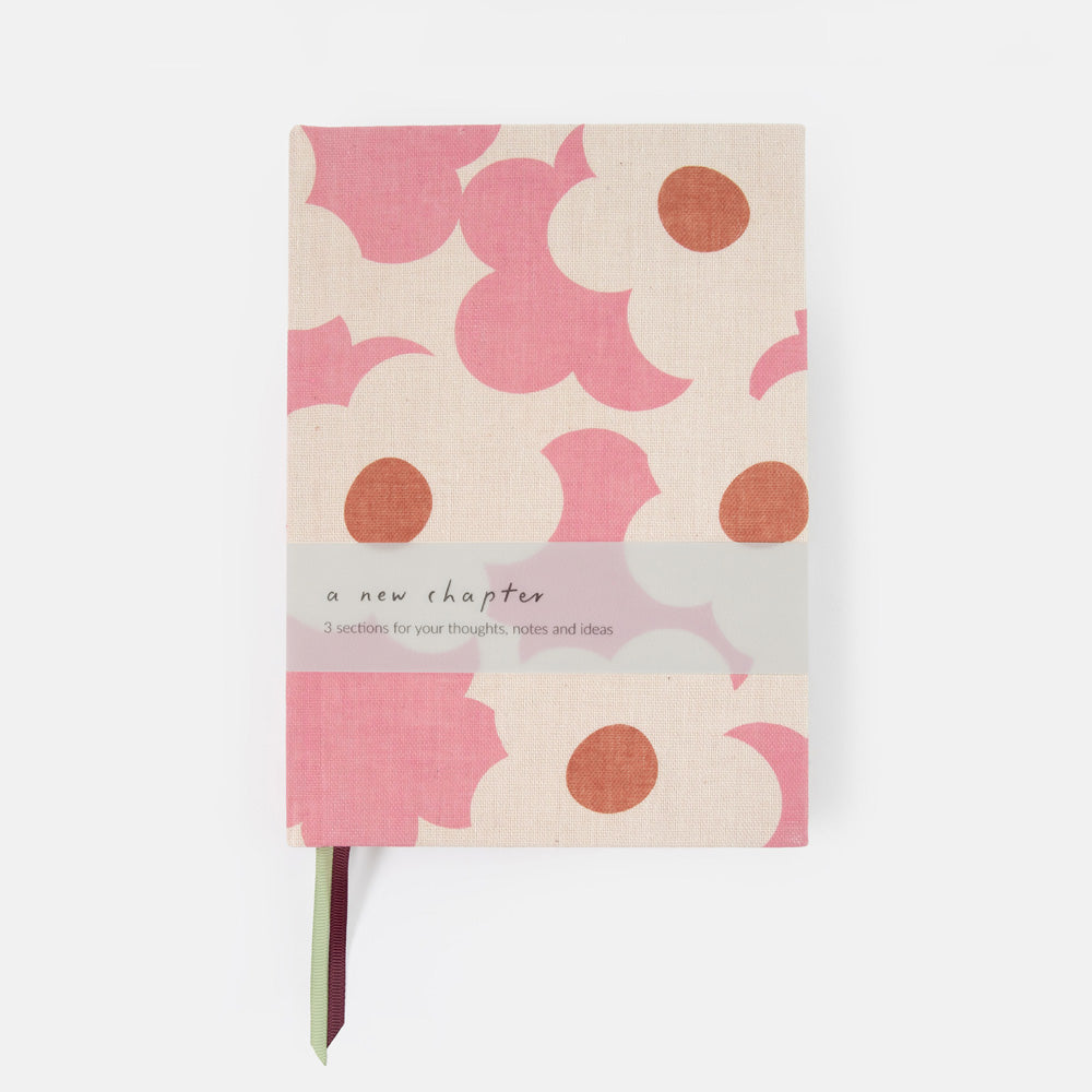 A hardcover notebook with a pink and beige abstract floral pattern, featuring a translucent paper band across the middle that reads 'a new chapter' and a thin ribbon bookmark peeking out from the bottom.