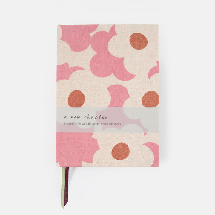A hardcover notebook with a pink and beige abstract floral pattern, featuring a translucent paper band across the middle that reads 'a new chapter' and a thin ribbon bookmark peeking out from the bottom.