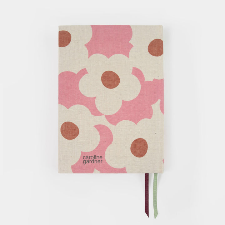 A Caroline Gardner notebook with a pink and cream floral pattern on a light beige linen cover, featuring a dark burgundy and light green ribbon bookmark.