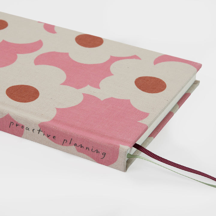 A hardcover planner or journal with a pink and cream floral pattern, featuring the words 'proactive planning' on the front cover and a dark red ribbon bookmark.