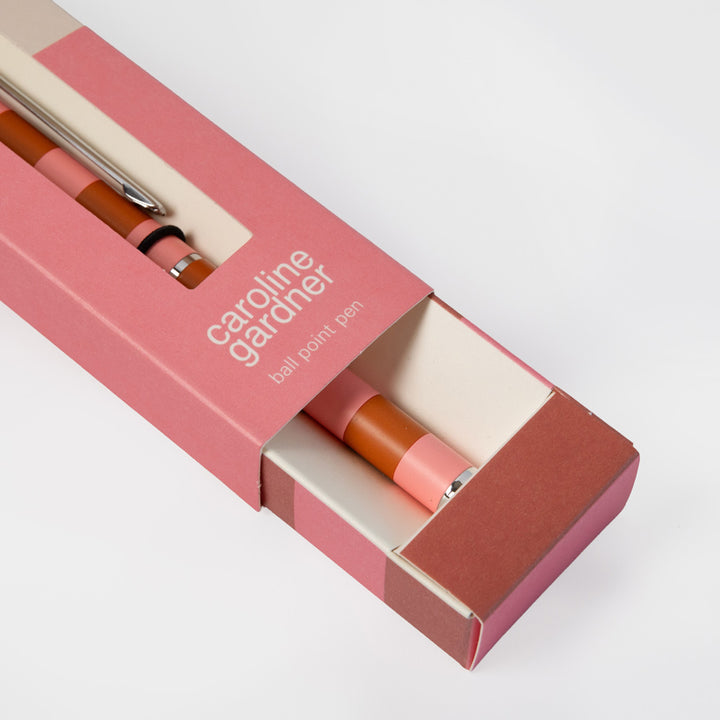 A Caroline Gardner ball point pen with a pink and orange body, partially visible inside its matching pink packaging box on a clean white background.