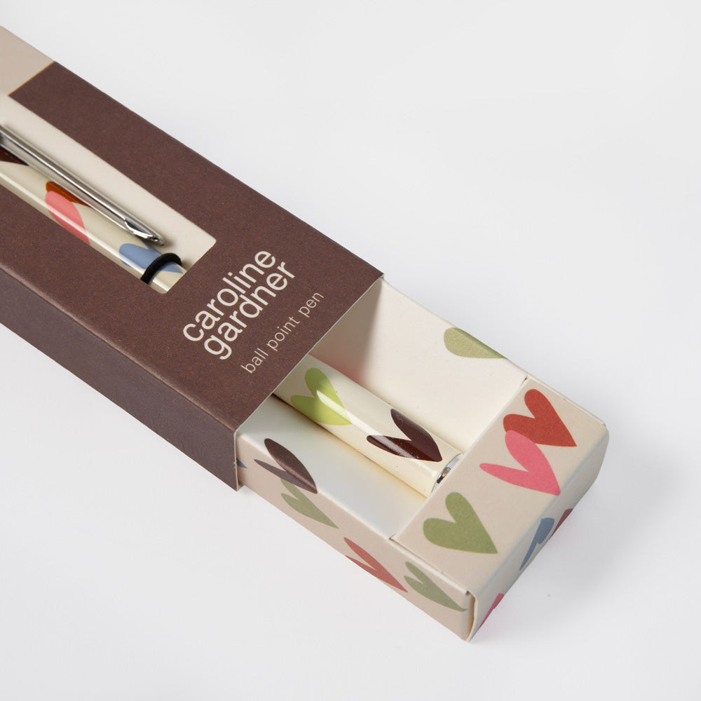 A Caroline Gardner ballpoint pen with a colorful heart pattern, shown partially inside its brown cardboard packaging box on a clean white background.