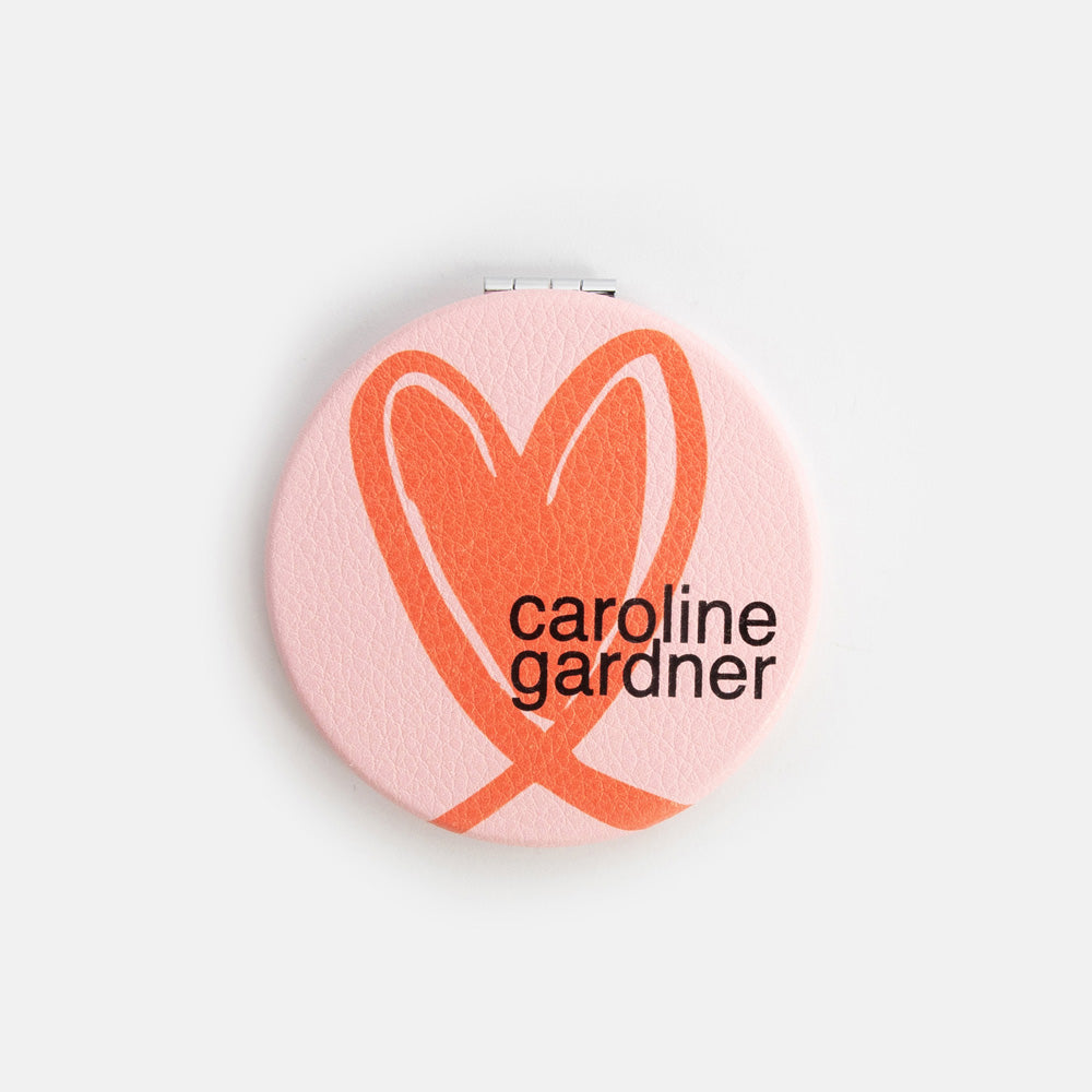 Compact mirror with pink background and orange heart design, featuring 'caroline gardner' branding.