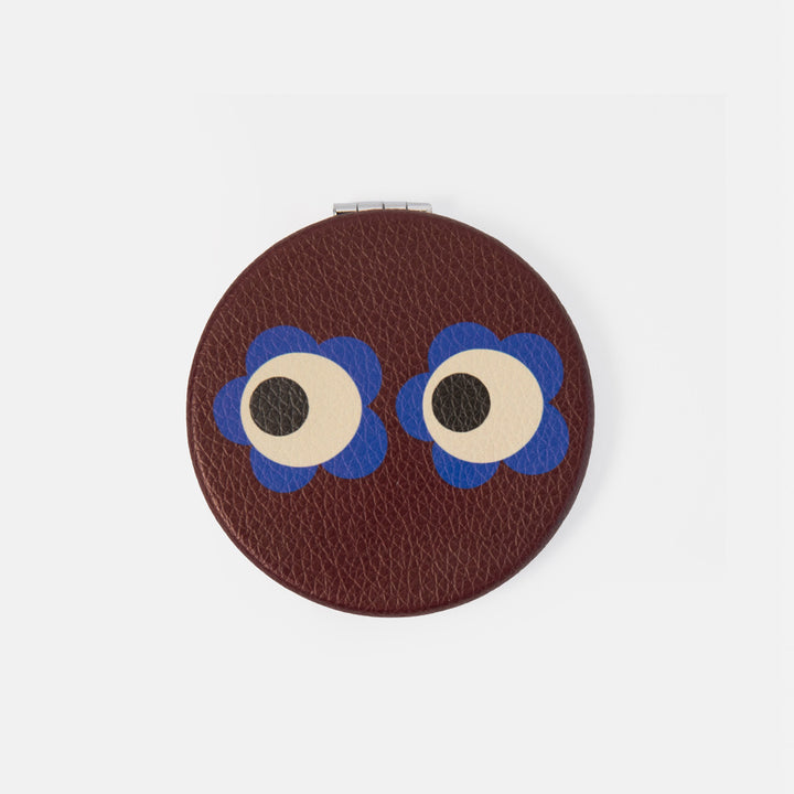 A round brown textured leather compact mirror featuring a playful face design with two large cartoon eyes that have blue flower-like outlines and cream-colored sclera, set against a plain light gray background.