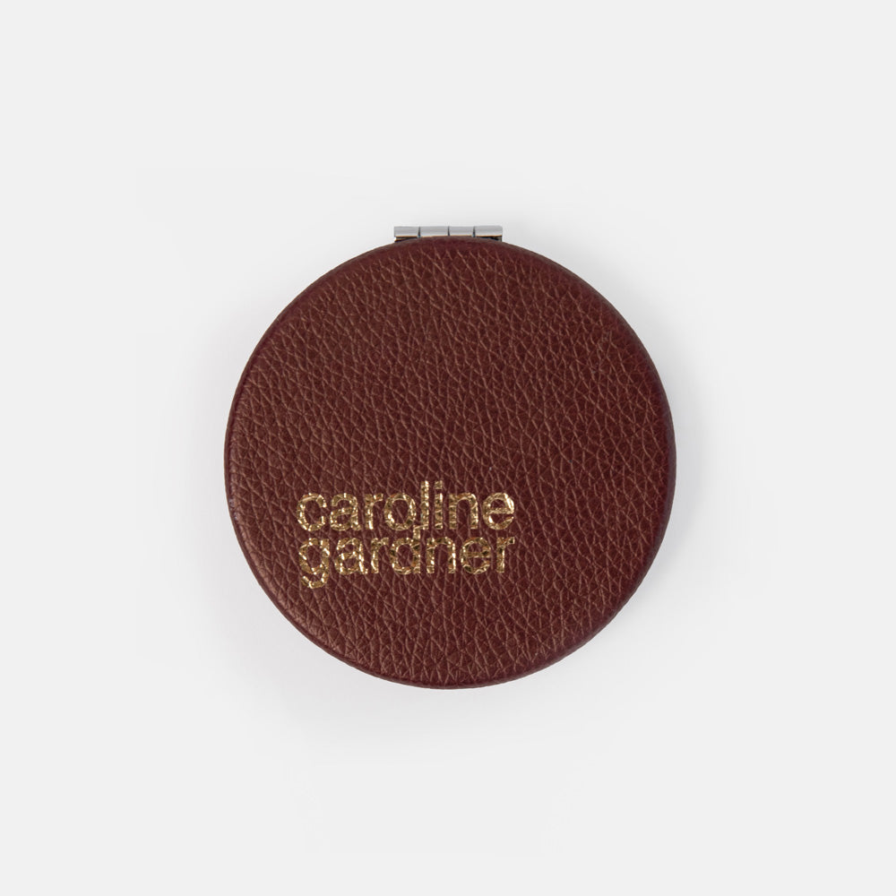 A round brown textured leather compact case with gold foil embossed branding reading 'caroline gardner' on a clean white background.
