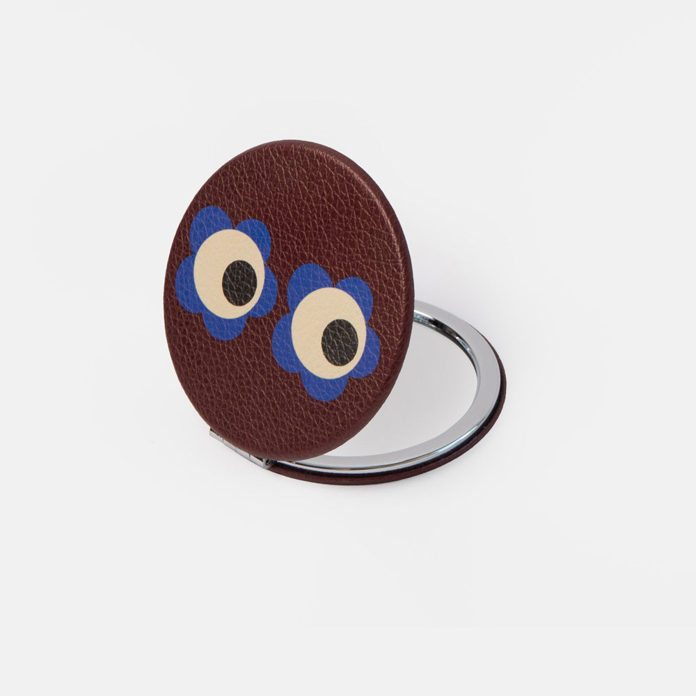 A round brown textured compact mirror with a silver metal stand, decorated with two cartoon eyes featuring blue petals and beige centers.