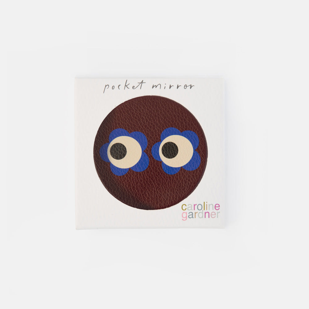 A square white product card featuring a circular brown textured design with cartoon eyes and blue flower-like accents, labeled 'pocket mirror' and 'caroline garçon'.