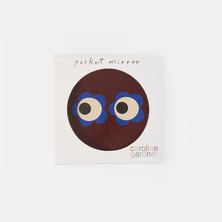 A square white product card featuring a circular brown textured design with cartoon eyes and blue flower-like accents, labeled 'pocket mirror' and 'caroline garçon'.