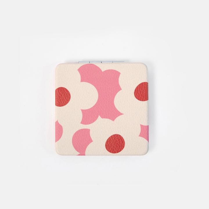 A square compact case with rounded corners, featuring a retro pattern of pink abstract shapes and red circles on a light beige textured background, set against a plain white studio background.