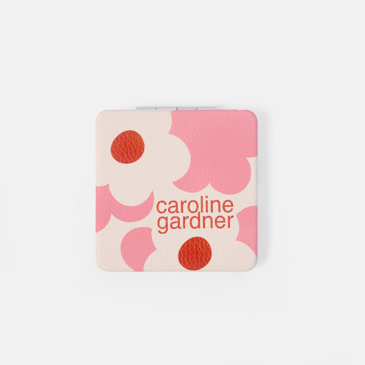 A compact mirror with a pink and white floral pattern and the brand name 'caroline gardner' printed in orange-red text, shown on a light gray background.