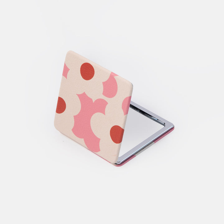 A small square compact mirror with a cream-colored cover decorated with pink and red abstract floral shapes, shown open on a light gray background.