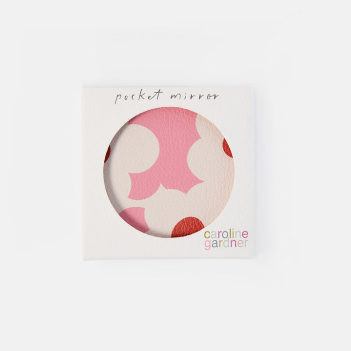A square white packaging card for a pocket mirror by Caroline Gardner, featuring a circular cutout that reveals a pink and cream patterned mirror surface.