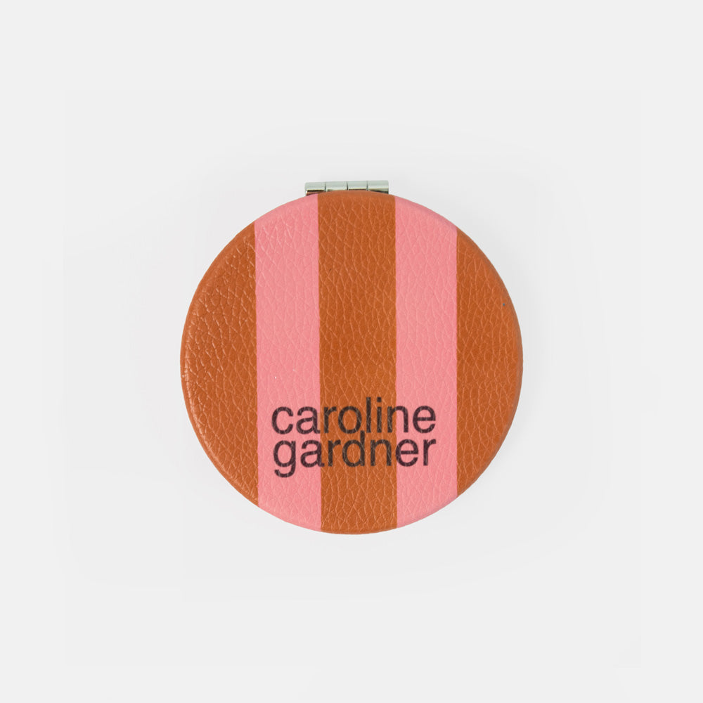 A round compact mirror with a textured leather-like finish featuring vertical stripes in terracotta brown and pink, with the brand name 'caroline gardner' printed in black text.