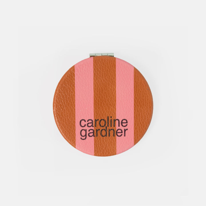 A round compact mirror with a textured leather-like finish featuring vertical stripes in terracotta brown and pink, with the brand name 'caroline gardner' printed in black text.