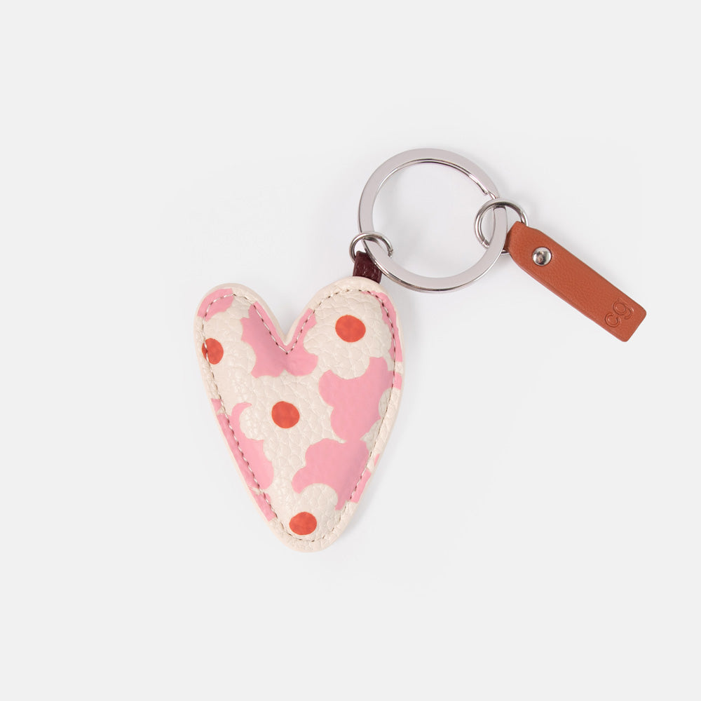 A heart-shaped keyring featuring a pink and cream floral pattern with red polka dots, attached to a silver metal key ring and a brown leather tag on a light gray background.