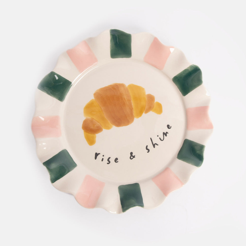 Ceramic plate with a croissant illustration and 'Rise & Shine' text on a white background