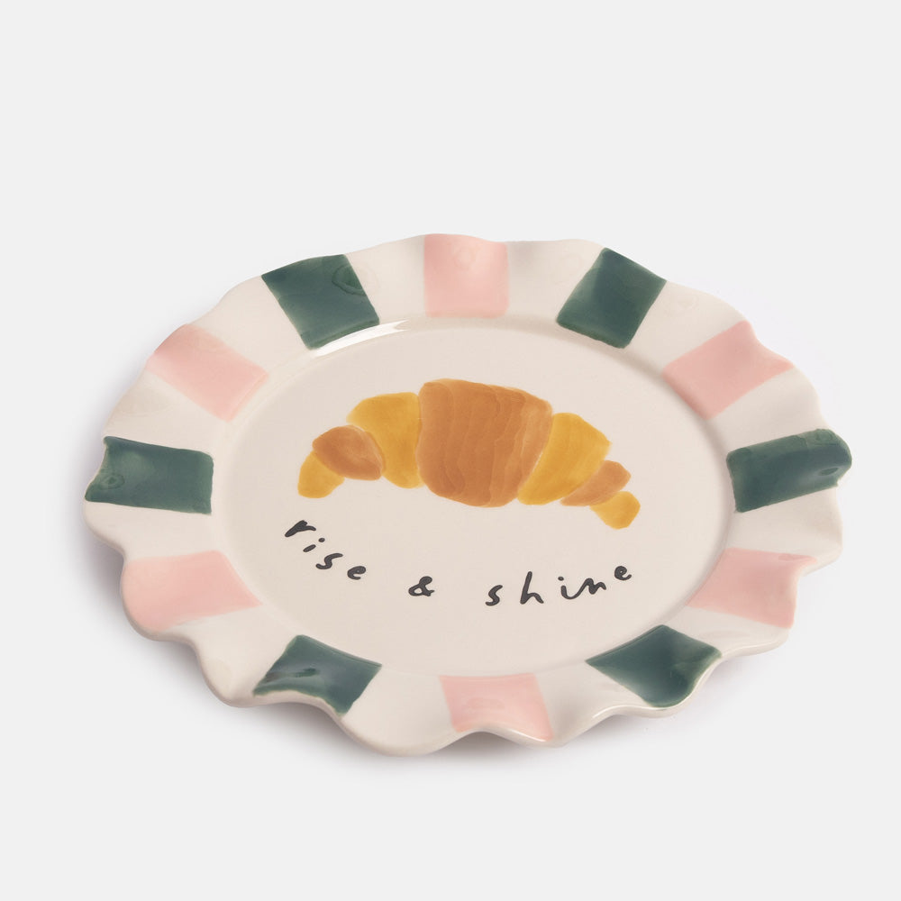Ceramic plate with a croissant illustration and 'Rise & Shine' text on a white background
