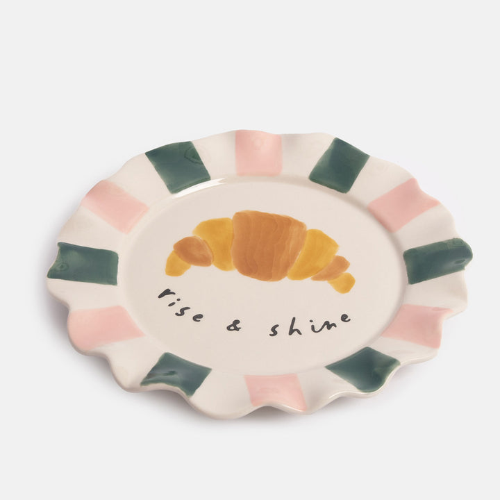 Ceramic plate with a croissant illustration and 'Rise & Shine' text on a white background