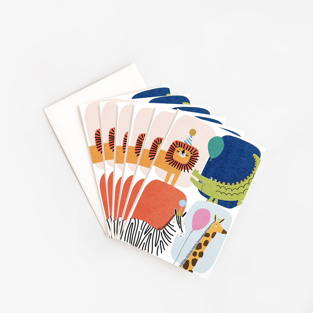 Six notecards fanned out next to a white envelope, each illustrated with a lion in a party hat, a green crocodile holding a balloon, a zebra and a giraffe with a pink balloon on blocks of pink, navy, coral and pale blue.