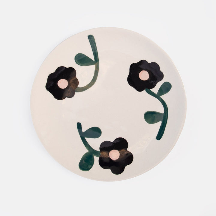 A round ceramic plate with a warm off-white background, decorated with three hand-painted black flowers and dark green stems.