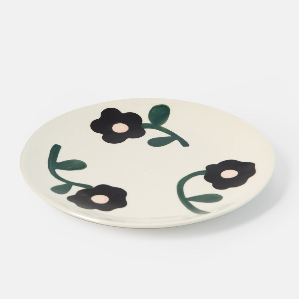 An off-white ceramic plate featuring a minimalist black flower and green vine pattern, photographed on a clean light gray background.
