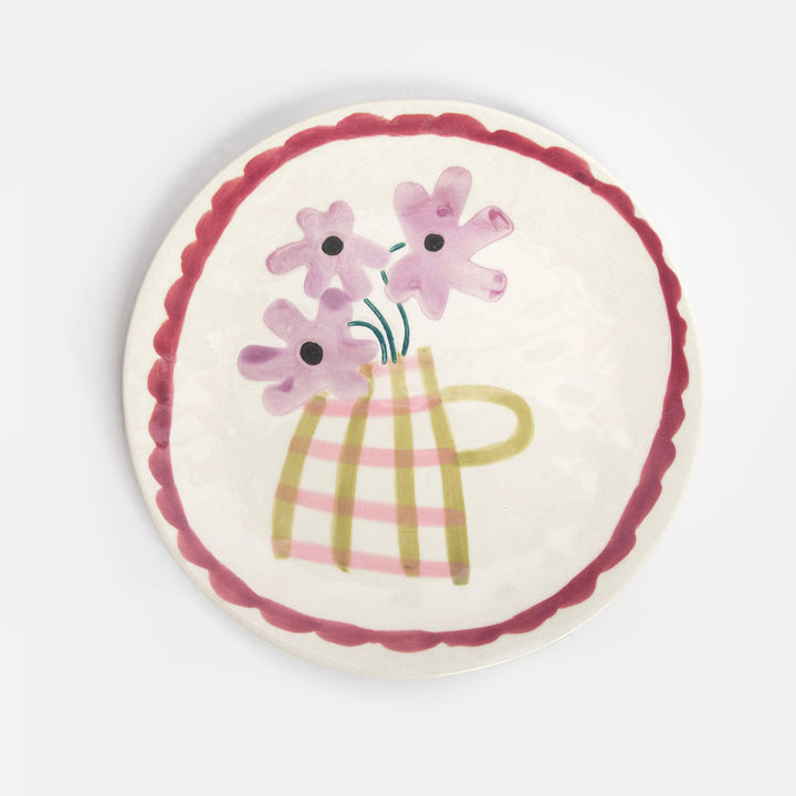 A round ceramic plate with a scalloped dark red rim, featuring a hand-painted illustration of pink flowers in a striped yellow-green vase on a cream background.