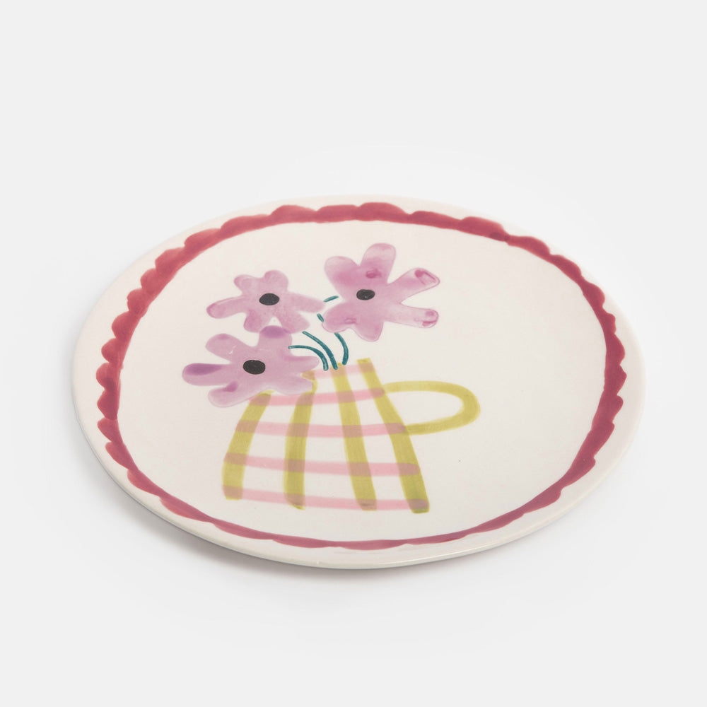 A round ceramic plate with a scalloped rim, featuring a hand-painted illustration of pink flowers in a yellow and pink plaid vase on a light cream background.