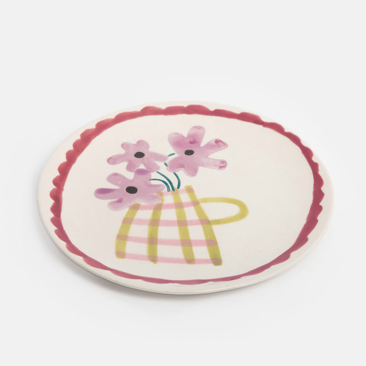 A round ceramic plate with a scalloped rim, featuring a hand-painted illustration of pink flowers in a yellow and pink plaid vase on a light cream background.