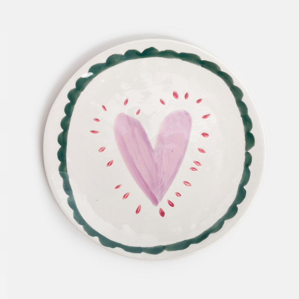 A round white ceramic plate with a hand-painted pink heart in the center, surrounded by small red dots, and a scalloped dark green border.