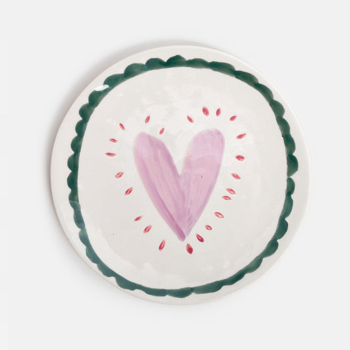 A round white ceramic plate with a hand-painted pink heart in the center, surrounded by small red dots, and a scalloped dark green border.