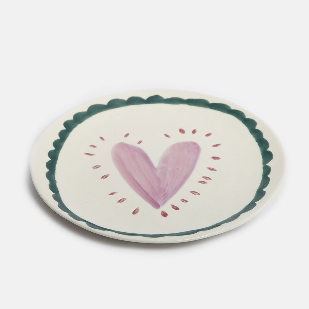 An round ceramic plate with a scalloped dark green border, featuring a hand-painted pink heart in the center surrounded by small pink dots, set against a plain light gray background.