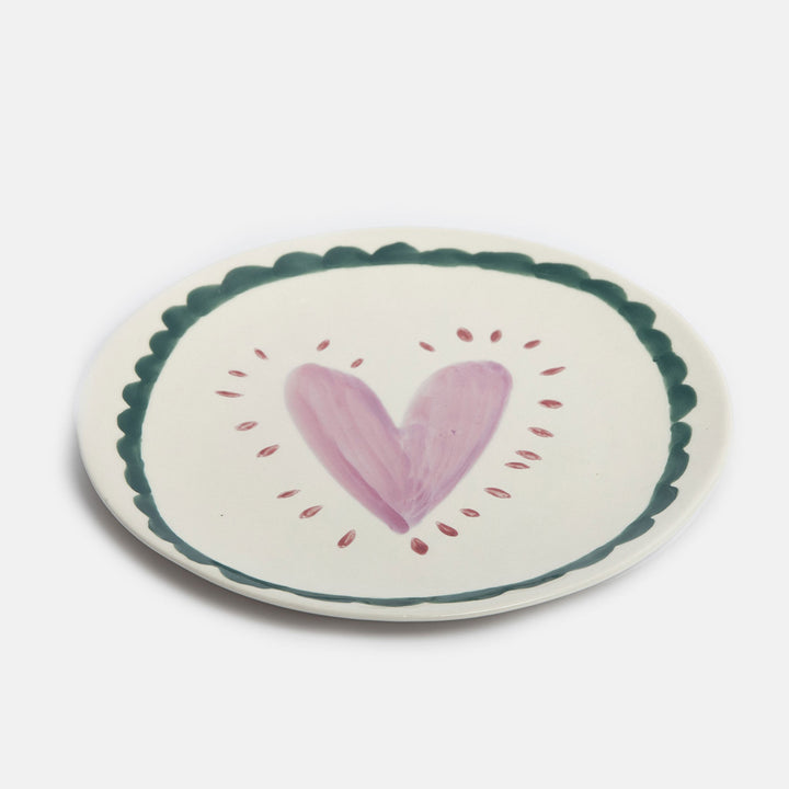 An round ceramic plate with a scalloped dark green border, featuring a hand-painted pink heart in the center surrounded by small pink dots, set against a plain light gray background.