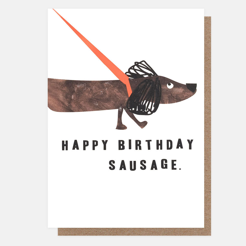 A birthday greeting card with a white front featuring a whimsical illustration of a dachshund wearing a black wire harness and an orange leash, with the text 'HAPPY BIRTHDAY SAUSAGE.' Below the card is a brown kraft envelope on a light gray background.