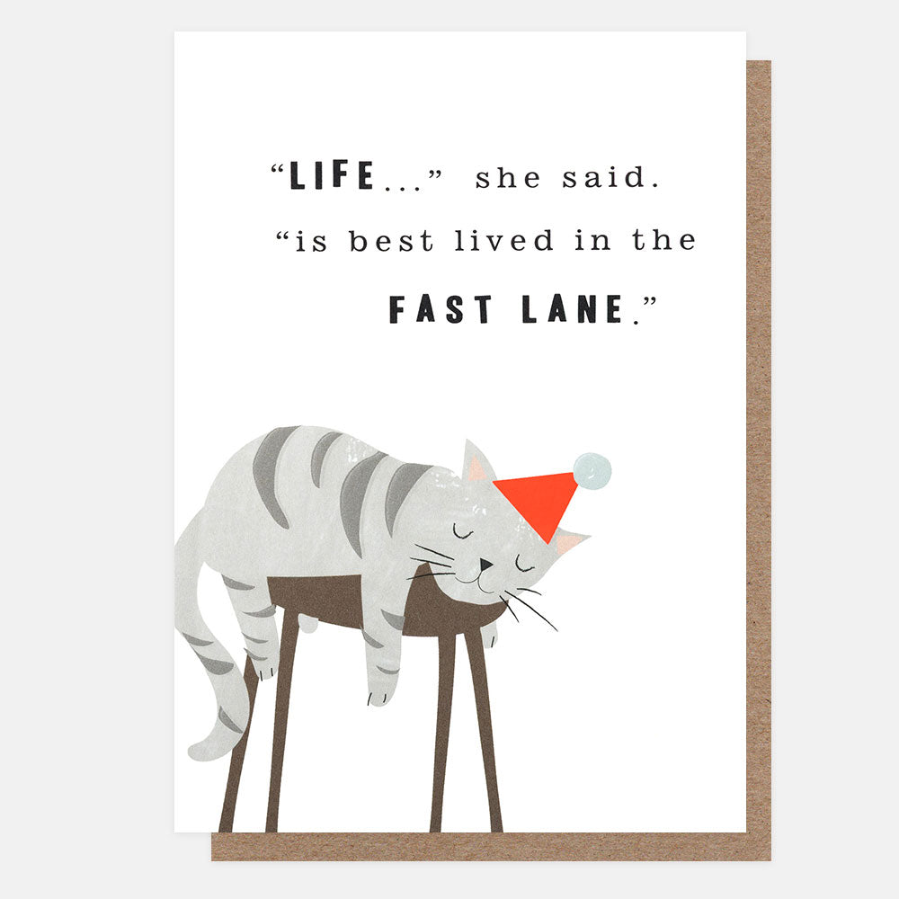 A greeting card with a brown kraft envelope behind it. The card features a sleeping grey cat on a stool wearing a party hat on a white background with a black quote that reads, "LIFE...she said. is best lived in the FAST LANE".