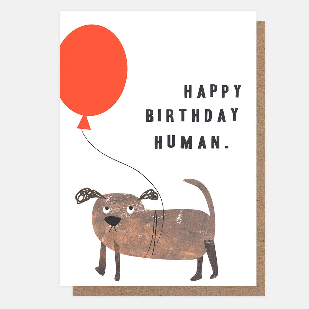 A greeting card with a brown dog illustration holding a red balloon and the text 'HAPPY BIRTHDAY HUMAN.' on a white card, with a brown kraft envelope behind it.