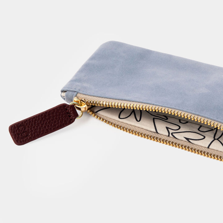 A close-up of a light blue suede pouch with a gold zipper, a gold-tone pull tab, and a beige interior printed with black graffiti-style lettering.
