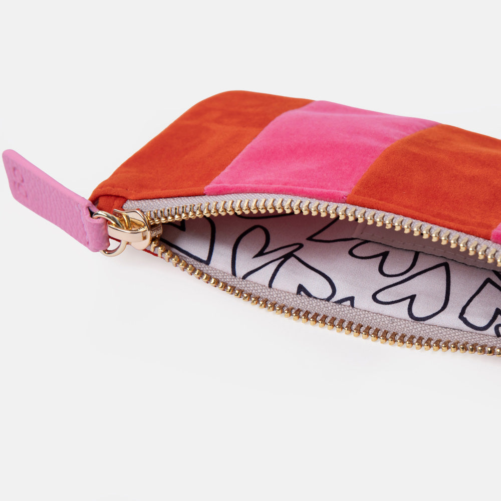 A close-up of a rectangular zip pouch with a patchwork design in orange, pink, and red velvet, shown open to reveal a white interior printed with black line art and a gold-tone zipper.