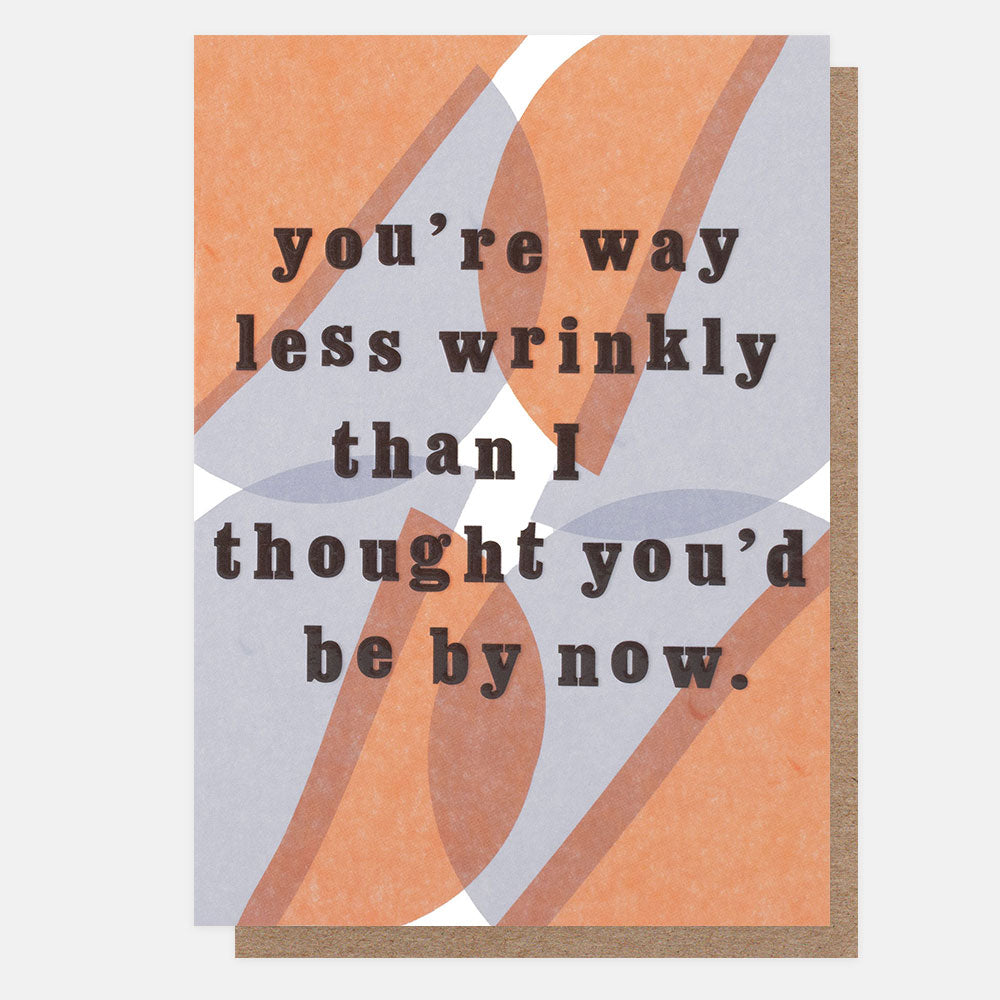 A square greeting card with a warm abstract design in terracotta, taupe, and light gray, featuring the text 'you're way less wrinkly than I thought you'd be by now.' in dark brown lettering, shown with a kraft envelope behind it on a light gray background.