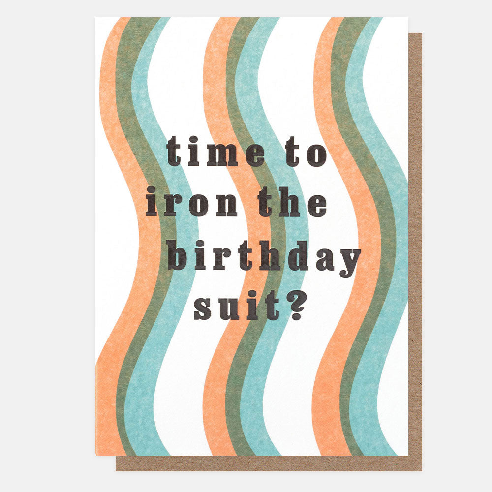 A greeting card with a brown envelope behind it, featuring wavy vertical stripes in peach, sage green, and light blue on a white background, with the text 'time to iron the birthday suit?' in dark gray lowercase letters.