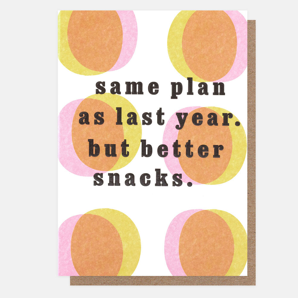 A greeting card with an orange, pink & yellow circle pattern and the text 'same plan as last year. but better snacks.' in dark gray, shown with a brown envelope behind it on a light gray background.