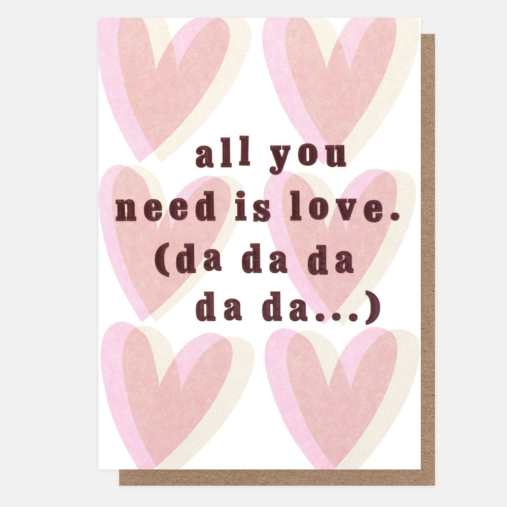 A greetings card with overlapping pink and beige hearts on a white background and the text 'all you need is love. (da da da da da...)' in plum embossed lettering, shown with a brown kraft envelope behind it.