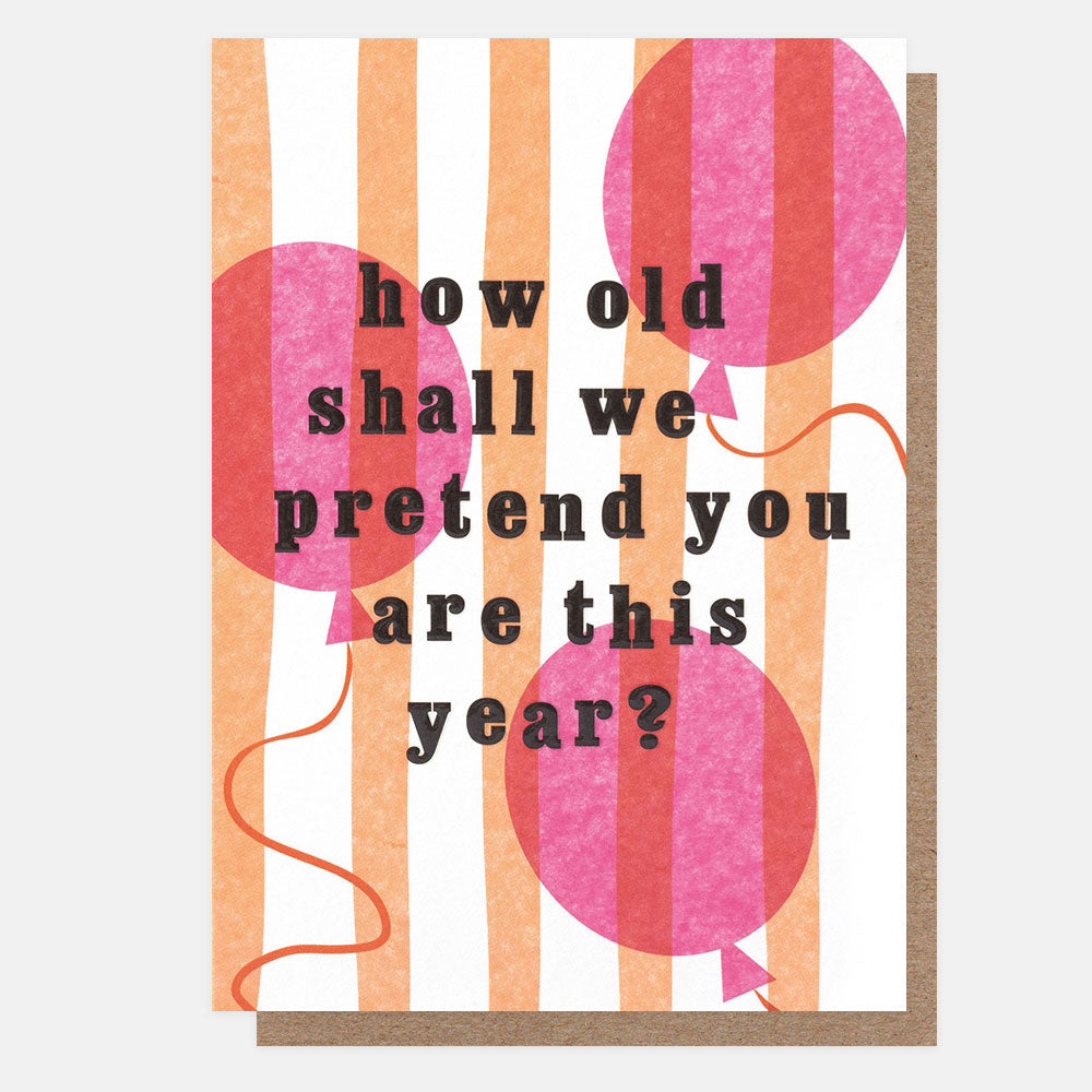 A greetings card with pink and red balloons on orange and white stripes and the text 'how old shall we pretend you are this year?' in black embossed lettering, shown with a brown kraft envelope behind it.