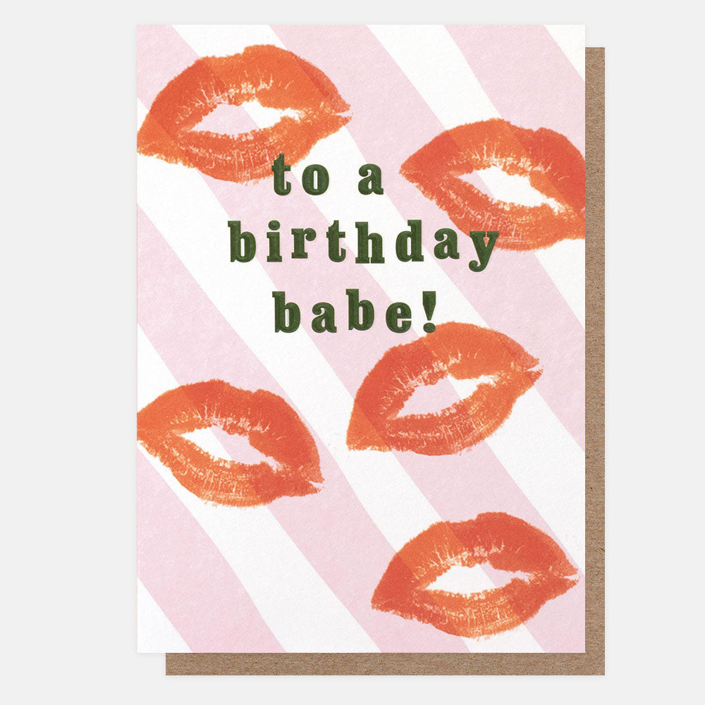 A greetings card with red lipstick kiss prints on pink and white diagonal stripes and the text 'to a birthday babe!' in green embossed lettering, shown with a brown kraft envelope behind it.