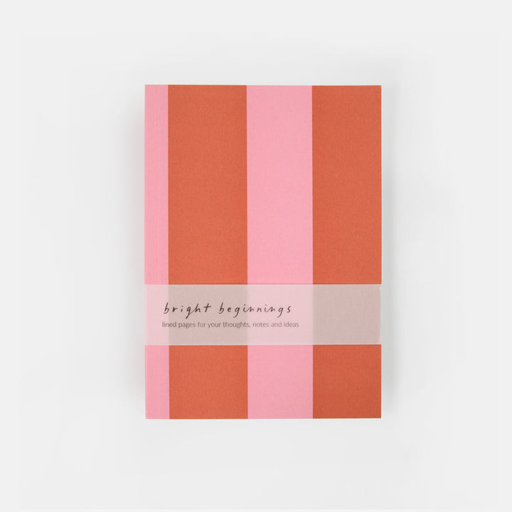A notebook with a striped cover in shades of pink and terracotta orange, wrapped with a white paper band that reads 'bright beginnings'.