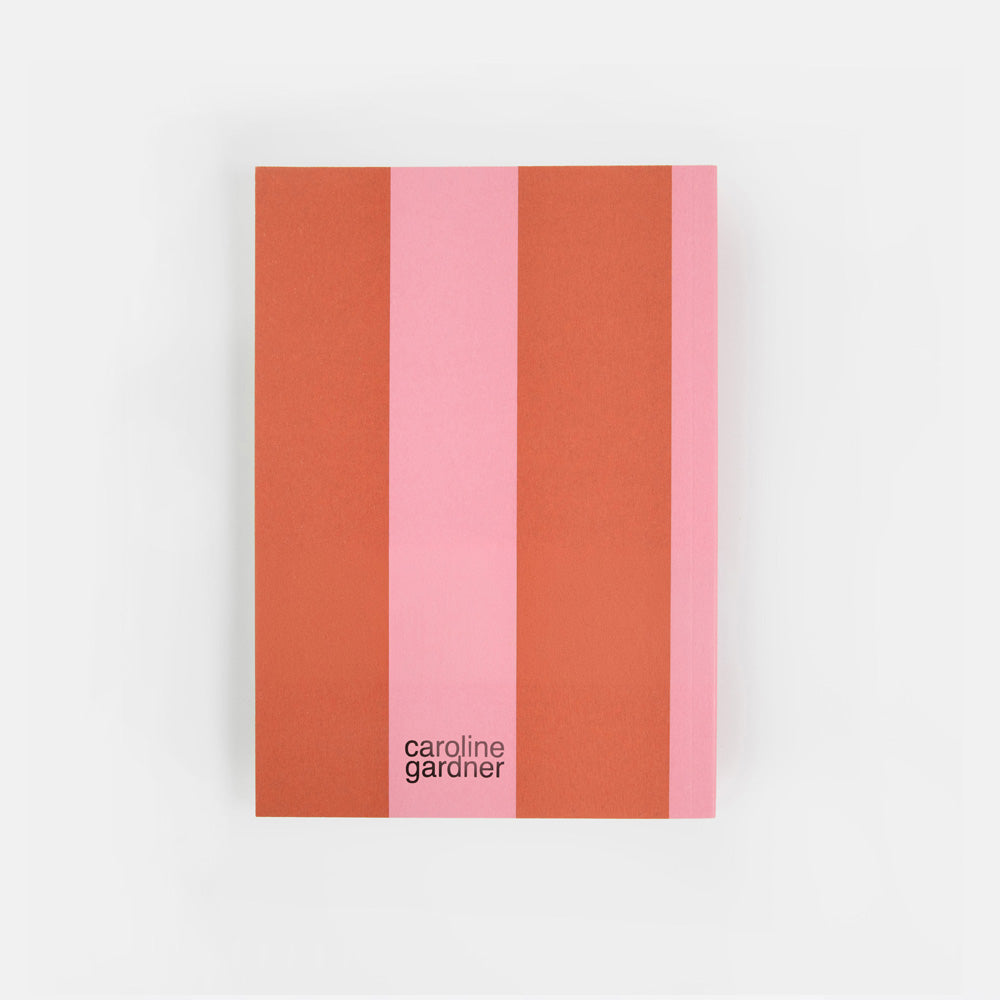 A notebook or journal with a minimalist cover design featuring vertical stripes in terracotta orange, light pink, and coral pink, with the text 'caroline garonier' printed in black near the bottom. The item is photographed flat against a clean, light gray background.