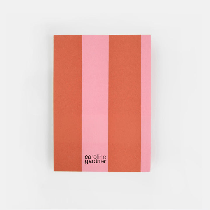 A notebook or journal with a minimalist cover design featuring vertical stripes in terracotta orange, light pink, and coral pink, with the text 'caroline garonier' printed in black near the bottom. The item is photographed flat against a clean, light gray background.