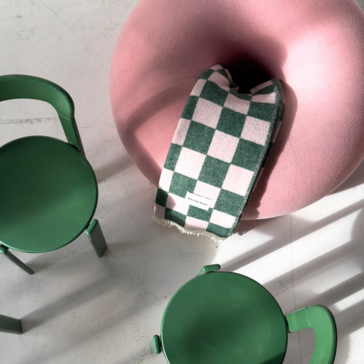 Pink and green checkerboard pattern pure wool blanket, woven in Europe and made by Maison Deux. Shown folded on a pink chair, next to two green chairs.