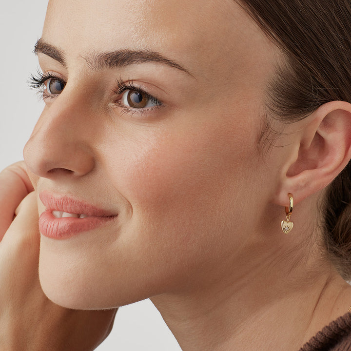 Close-up of a woman wearing a gold hoop earring with a heart charm on a neutral background